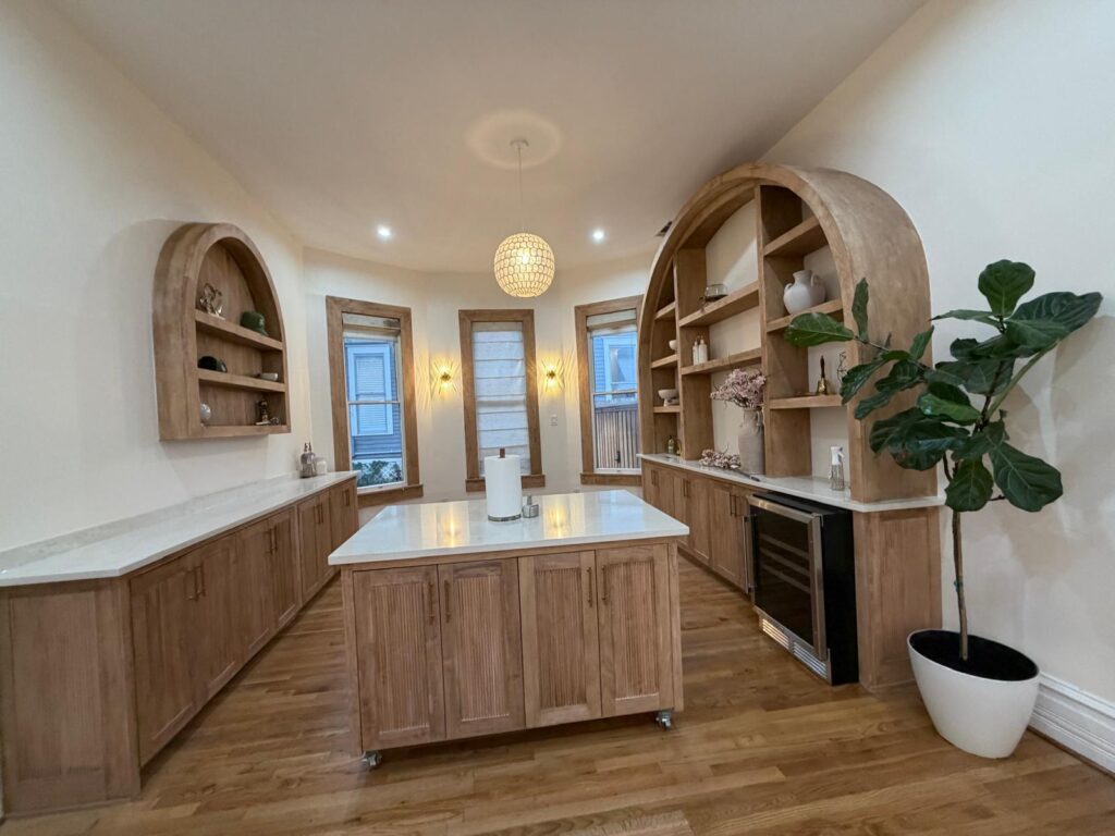Bright rustic kitchen with curved wooden shelves, central island, and a tall potted plant by the windowed wall.