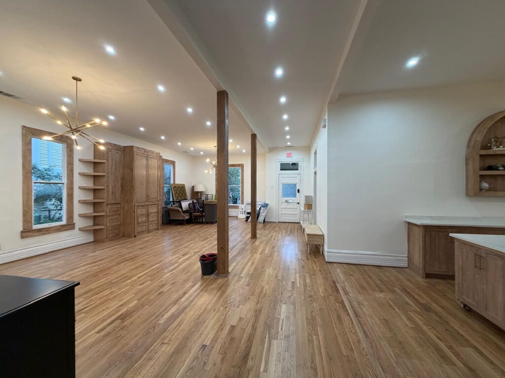 Spacious showroom with wood flooring, built-in cabinets, and tall wooden posts; modern pendant lights and a doorway at the back center.